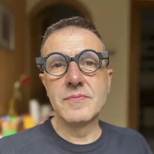 A man wearing glasses and a blue blazer, with short brown hair, looks at the camera. A stone wall is in the background.